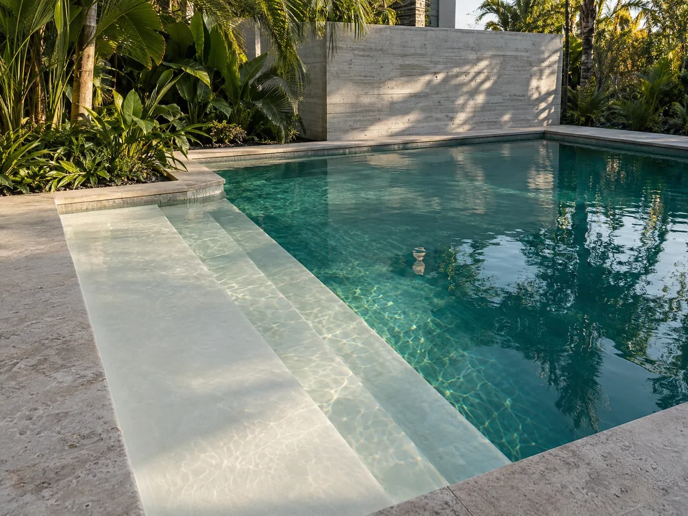 A pool with a wide sun shelf and bench step in bright white plaster, beside travertine coping and planting